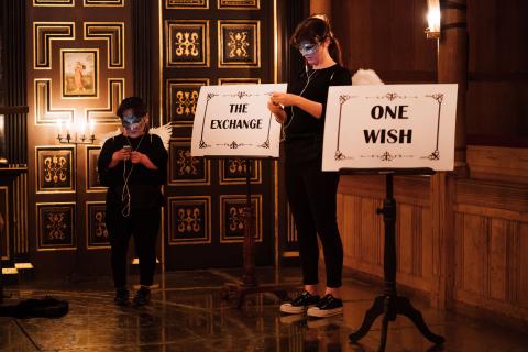 Lisa and Rachael dress in black with angel wings and glittery eye masks looking at phones with headphones and two signs saying THE EXCHANGE and ONE WISH